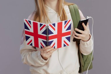 Sırt çantalı kıza yakın çekim, İngilizce bayrak çalışma kitabı, kulaklık ve akıllı telefon griye karşı.