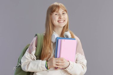 Sırt çantası, çalışma kitabı ve kulaklığı gri renge karşı olan bej eşofmanlı mutlu modern kız portresi.