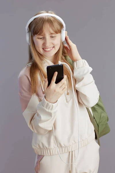 幸せなトレンディーな女の子でベージュトラックスーツでバックパックと音楽を聞いてヘッドフォンとグレーの背景に隔離されたスマートフォンアプリケーションを使用して — ストック写真