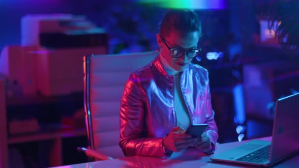 霓虹灯元的未来主义概念 现代40岁的女人 戴着眼镜 办公室里有笔记本电脑和智能手机 — 图库视频影像