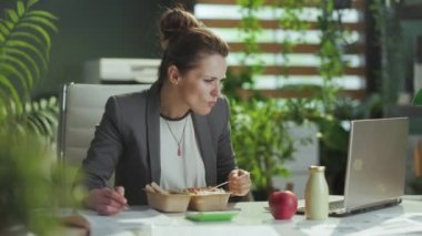 Sürdürülebilir iş yeri. Modern yeşil ofisteki gri takım elbiseli modern iş kadını. Laptop yiyor, salata yiyor ve akıllı telefon kullanıyor..