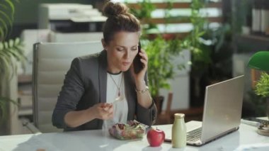 Sürdürülebilir iş yeri. Modern yeşil ofisteki gri takım elbiseli, orta yaşlı, gülümseyen bir kadın işçi. Laptop yiyor ve akıllı telefondan konuşuyor..