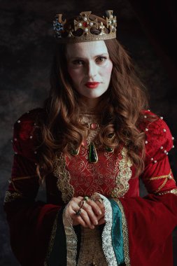Kırmızı elbiseli, beyaz makyajlı ve tacı koyu gri arka planda olan ortaçağ kraliçesi..