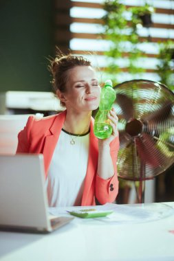 Sürdürülebilir iş yeri. Modern yeşil ofiste kırmızı ceketli, su şişeli ve elektrikli vantilatörlü mutlu modern kadın işçi..