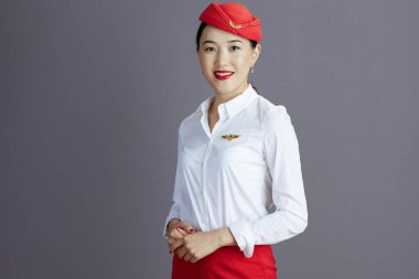 Gri arka planda izole edilmiş kırmızı etekli ve şapka formalı gülümseyen Asyalı bayan hostes..