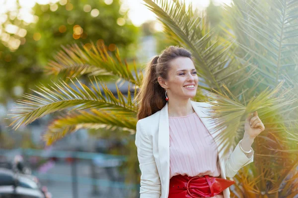 ピンクのドレスを着た幸せな現代女性と熱帯の木の近くの街の白いジャケット — ストック写真