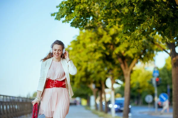 ピンクのドレスを着た流行の女性とヘッドフォンで音楽を聞いて街の白いジャケットを笑って歩く — ストック写真