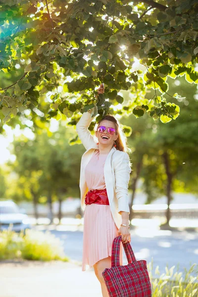 城市里穿着粉色衣服和白色夹克 头戴红包和太阳镜的时髦女人微笑着 — 图库照片