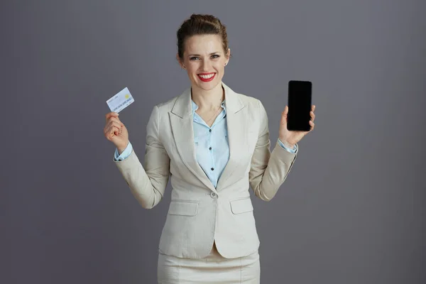スマートフォンやクレジットカードでグレーを背景にしたライトビジネススーツを着た現代女性従業員の笑顔 — ストック写真