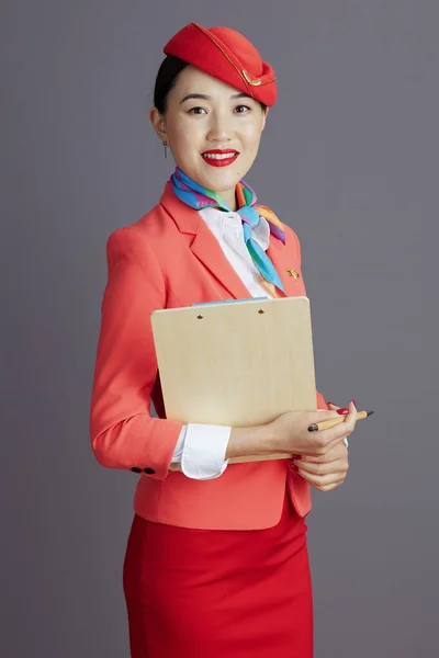 穿着红色短裙 夹克和帽子制服 戴着灰色剪贴板的现代亚洲女空姐微笑着 — 图库照片