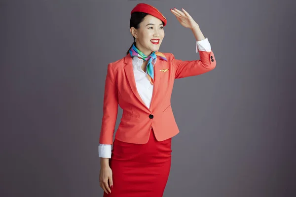 穿着红色短裙 夹克和帽子制服的快乐的现代亚洲女性空姐 凝视着被灰色隔离的远方 — 图库照片