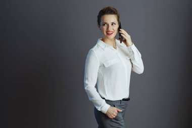 Mutlu, şık, küçük işletme sahibi beyaz bluzlu bir kadın cep telefonuyla konuşuyor ve gri arka planda izole edilmiş bir kenara bakıyor..