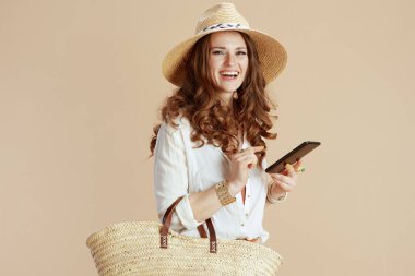 Plaj tatili Beyaz bluz ve şortla saman çantalı, yaz şapkalı, akıllı telefon kullanan mutlu stil sahibi kadın..