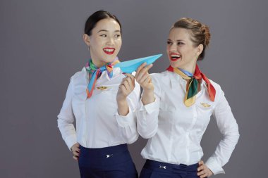 Gülümseyen zarif bayan hostesler mavi etekli, beyaz gömlekli ve atkılı gri arka planda kağıt uçakla.
