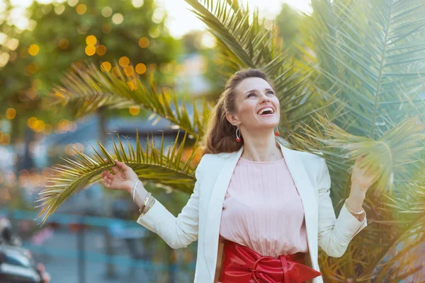 幸せな中年齢女性でピンクのドレスと白いジャケットで街の近く熱帯の木 — ストック写真