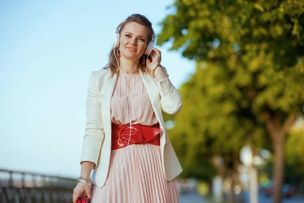 ピンクのドレスを着たスタイリッシュな女性とヘッドフォンで音楽を聴き歩く街の白いジャケット — ストック写真