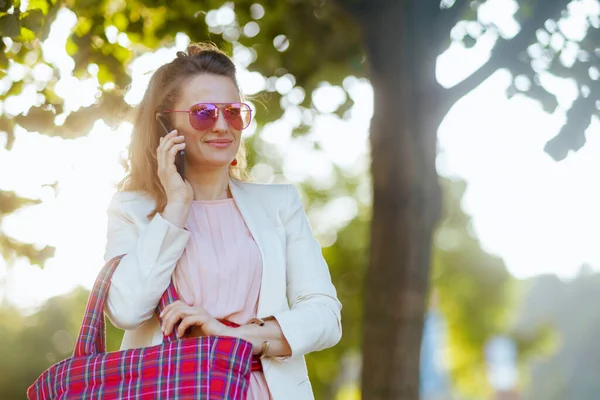 城市里穿着粉色衣服和白色夹克 头戴红包和太阳镜 头戴智能手机的优雅中年妇女 — 图库照片
