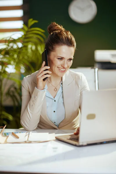 在现代绿色办公室穿着轻便西装 手持笔记本电脑 用智能手机聊天的快乐的现代中年妇女会计 — 图库照片