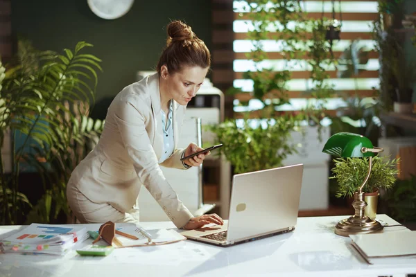 現代のグリーンオフィスのライトビジネススーツを着た中年女性従業員スマートフォンを使ったノートパソコンで — ストック写真