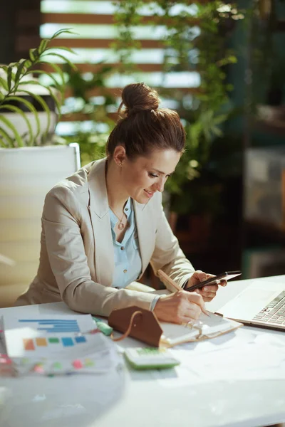 幸せな現代40歳の女性スマートフォンやノートブック付きのモダンな緑のオフィスで軽いビジネススーツの従業員 — ストック写真