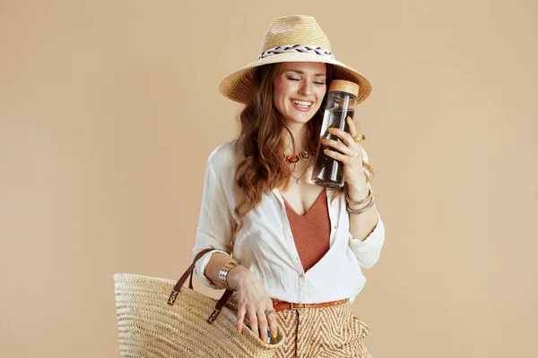 海滩度假 穿着白衬衫 头戴米黄色背景 头戴瓶装水 草袋和草帽的时髦中年女性微笑着 — 图库照片