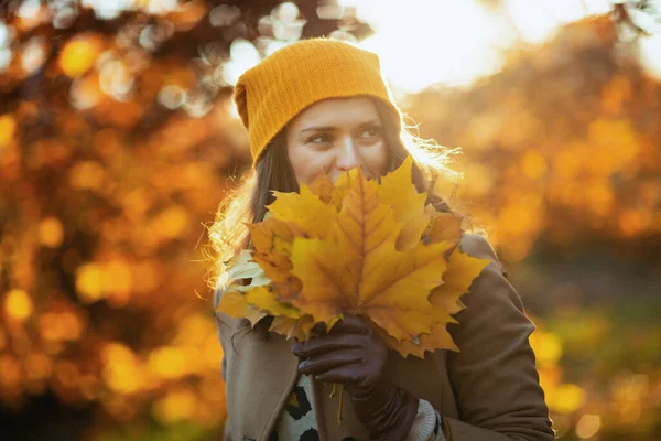 Hello autumn. stylish 40 years old woman in brown coat and yellow hat with autumn yellow leaves outside on the city park in autumn.