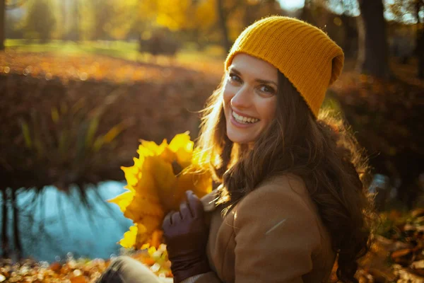 11月だ 幸せなトレンディーな40歳の女性茶色のコートと黄色の帽子秋に街の公園で屋外に座って黄色の葉 — ストック写真