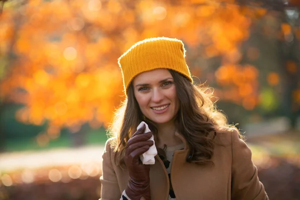 9月だ ベージュのコートを着たスタイリッシュな女性と秋に街の公園でナプキンが鼻を吹いてオレンジの帽子 — ストック写真