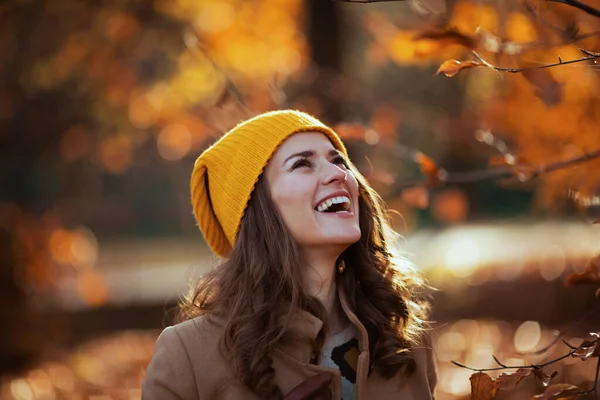 城市公园外身穿米黄色外套 头戴橙色帽子的优雅女性微笑着 — 图库照片
