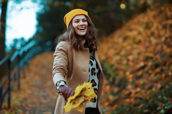 秋には公園の外で遊歩道を楽しむ黄色の葉を持つベージュコートとオレンジの帽子の幸せなスタイリッシュな女性 — ストック写真