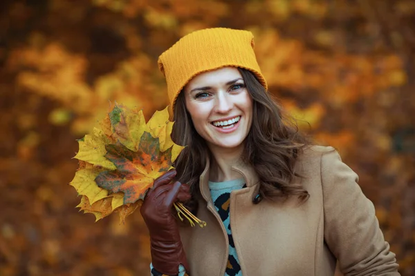 十一月 城市公园外 身穿米黄色外套 头戴橙色帽子 头戴秋天黄色叶子的中年妇女面带笑容 — 图库照片