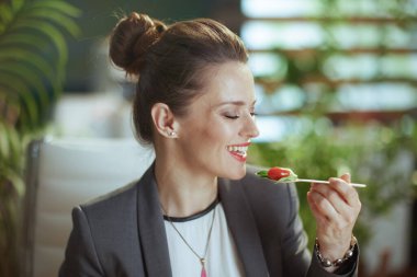 Sürdürülebilir iş yeri. Gülümseyen, modern, küçük işletme sahibi, gri takım elbiseli, yeşil ofiste salata yiyen bir kadın..