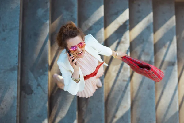城市里穿着粉色衣服和白色夹克 头戴红包和太阳镜 头戴智能手机 面带微笑的女人的上图 — 图库照片