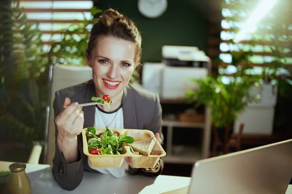 持続可能な職場 幸せな現代40歳の女性従業員グレービジネススーツで現代緑のオフィスでラップトップ食べてサラダ — ストック写真
