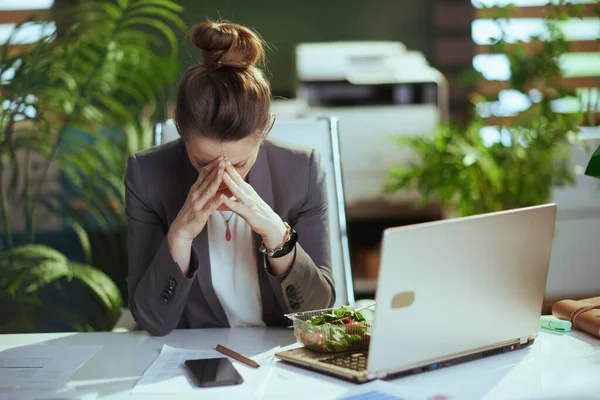 持続可能な職場 現代的な中年のブックキーパーの女性を強調グレーのビジネススーツで現代緑のオフィスでラップトップとスマートフォンがサラダを食べる — ストック写真
