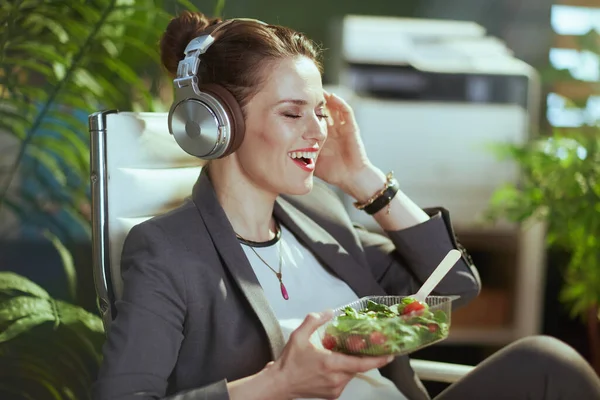 持続可能な職場 幸せな現代的な中年会計士女性で灰色のビジネススーツで現代緑のオフィスでヘッドフォンを食べサラダ — ストック写真