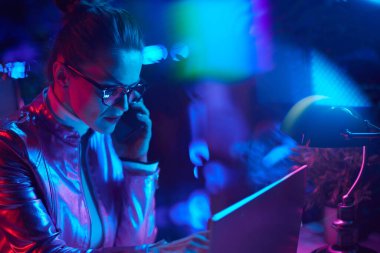 Neon metaevren fütüristik konsepti. Düşünceli, gözlüklü, dizüstü bilgisayar kullanan ve modern ofiste akıllı telefon kullanan zarif bir kadın..
