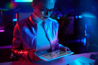 Neon metaevren fütüristik konsepti. Tabletli gözlüklü şık bir kadın modern ofiste grafiklerle çalışıyor..