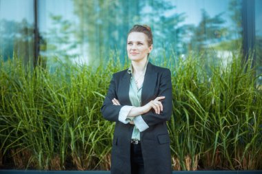 Çapraz kolları olan siyah ceketli, ofis binasının yanındaki düşünceli, orta yaşlı kadın işçi..