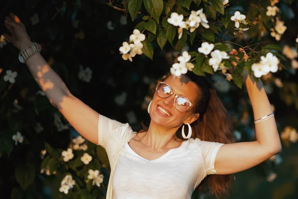 夏天的时候笑容可亲的年轻女性 身穿白衬衫 眼镜紧盯着开花的树 — 图库照片