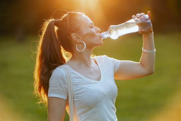 夏天的时候笑着穿白衬衫的现代女人在外面的草地上喝水 — 图库照片