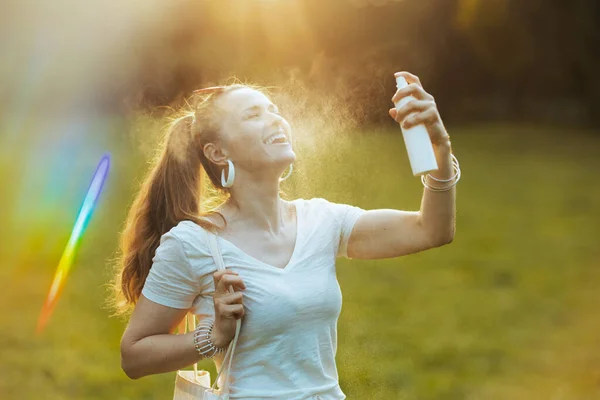 夏天的时候40岁穿着白衬衫的快乐而时尚的女人在外面的草地上使用面部喷雾 — 图库照片