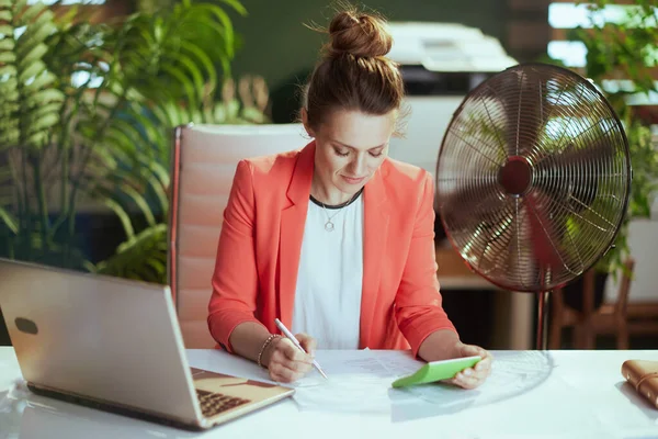 持続可能な職場 現代40歳の中小企業経営者の女性は 電気ファンとラップトップで赤いジャケットで仕事をしています — ストック写真