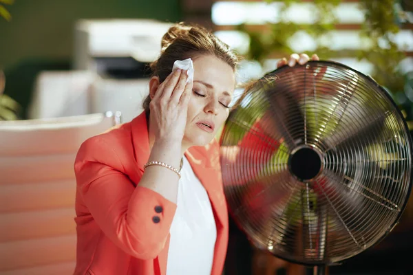 持続可能な職場 現代的な中年中小企業所有者女性で現代緑のオフィスで赤いジャケットで電気ファンと夏の暑さに苦しんでナプキン — ストック写真