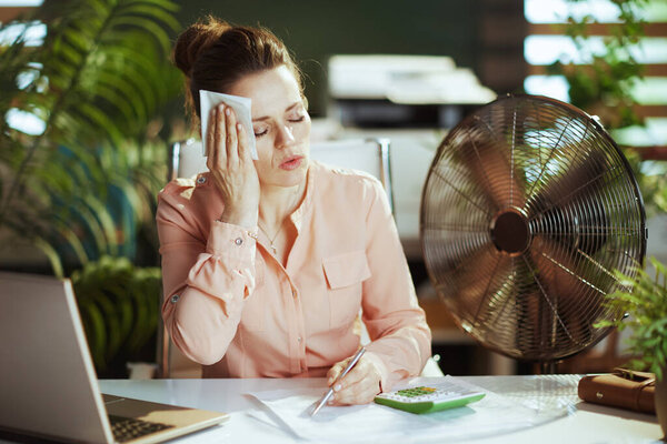 Sustainable workplace. modern 40 years old woman worker at work with electric fan, laptop and napkin suffering from summer heat.