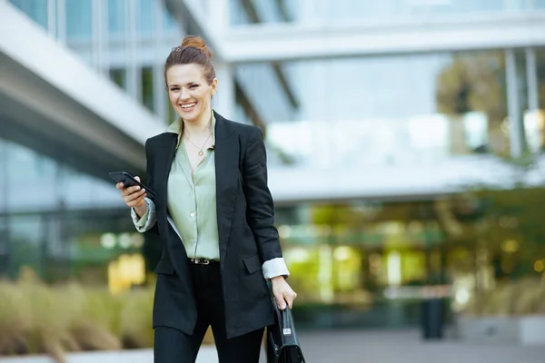 smiling modern middle aged woman worker near business center in black jacket with briefcase using smartphone and walking.