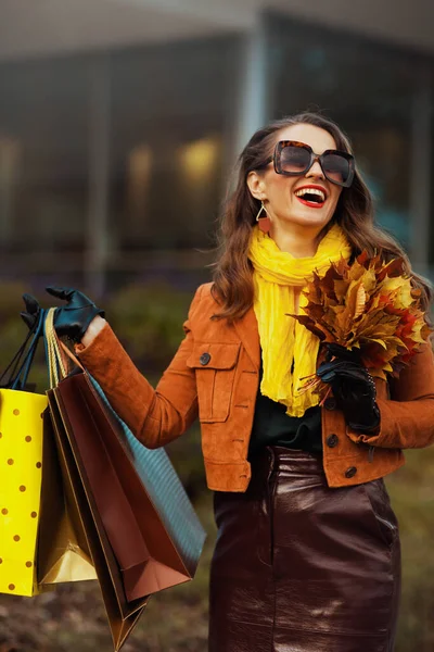 11月だ ショッピングバッグと秋の黄色の葉を持つ茶色のトレンチコートで流行の女性を笑顔街の店の近く — ストック写真