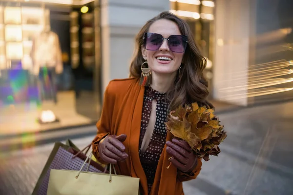 城市里 穿着橙色风衣 提着购物袋和秋天黄色叶子的中年妇女在商店附近快乐地生活着 — 图库照片