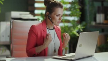 Sürdürülebilir iş yeri. Modern yeşil ofisteki kızgın modern iş kadını kırmızı ceketli, kulaklı ve dizüstü bilgisayarlı..