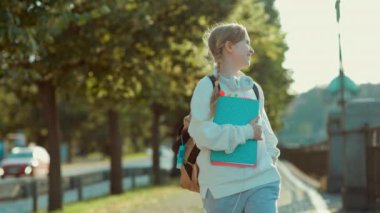 smiling stylish girl in white sweatshirt with backpack and headphones going to school outdoors in the city.